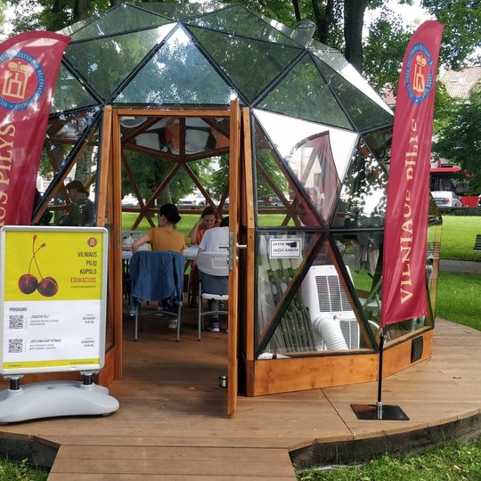 Vilniaus pilių kupolas su vidujė esančiais žmonėmis ir atviromis durimis. Šalia įėjimo Vilniaus pilių vėliavos, kairėje renginio plakatas.