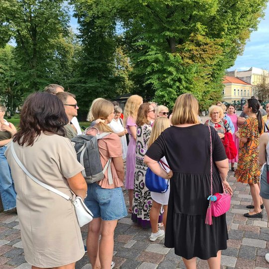 Įvairaus amžiaus eksursijos dalyviai klausosi gidės pasakojimų Katedros aikštėje.