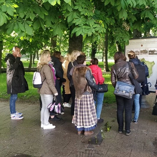 Žmonės vaikšto po pilių parką ir klausosi ekskursijos. 