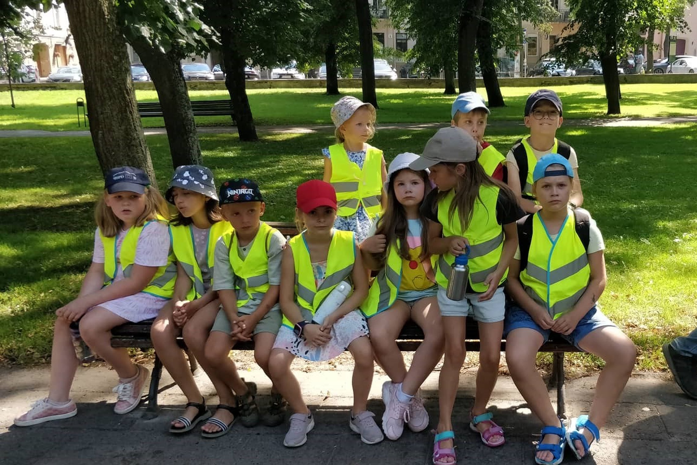 Vaikai su atšvaitinėmis liemenėmis sėdi amt suoliuko parkelyje šalai Vilniaus pilių kupolo.
