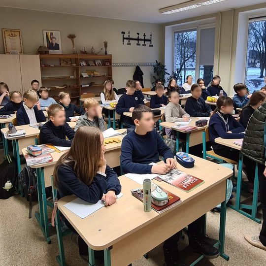 Klasė, pilna mokinių. Mokytoja stovi priekyje. Vaikai ruošiasi gaminti savo herbus.