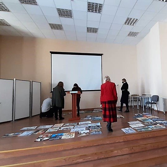 Erdvios salės fragmentas: trys ištisiniai laipteliai ir blizgios rudos grindys, ant priešakinės sienos – baltas ekranas, dešinėje prie sienos - stalai, kėdės, pakabinamos lubos su įmontuotais keturkampiais šviestuvais, viduryje – ant laibo stovo tribūnėlė