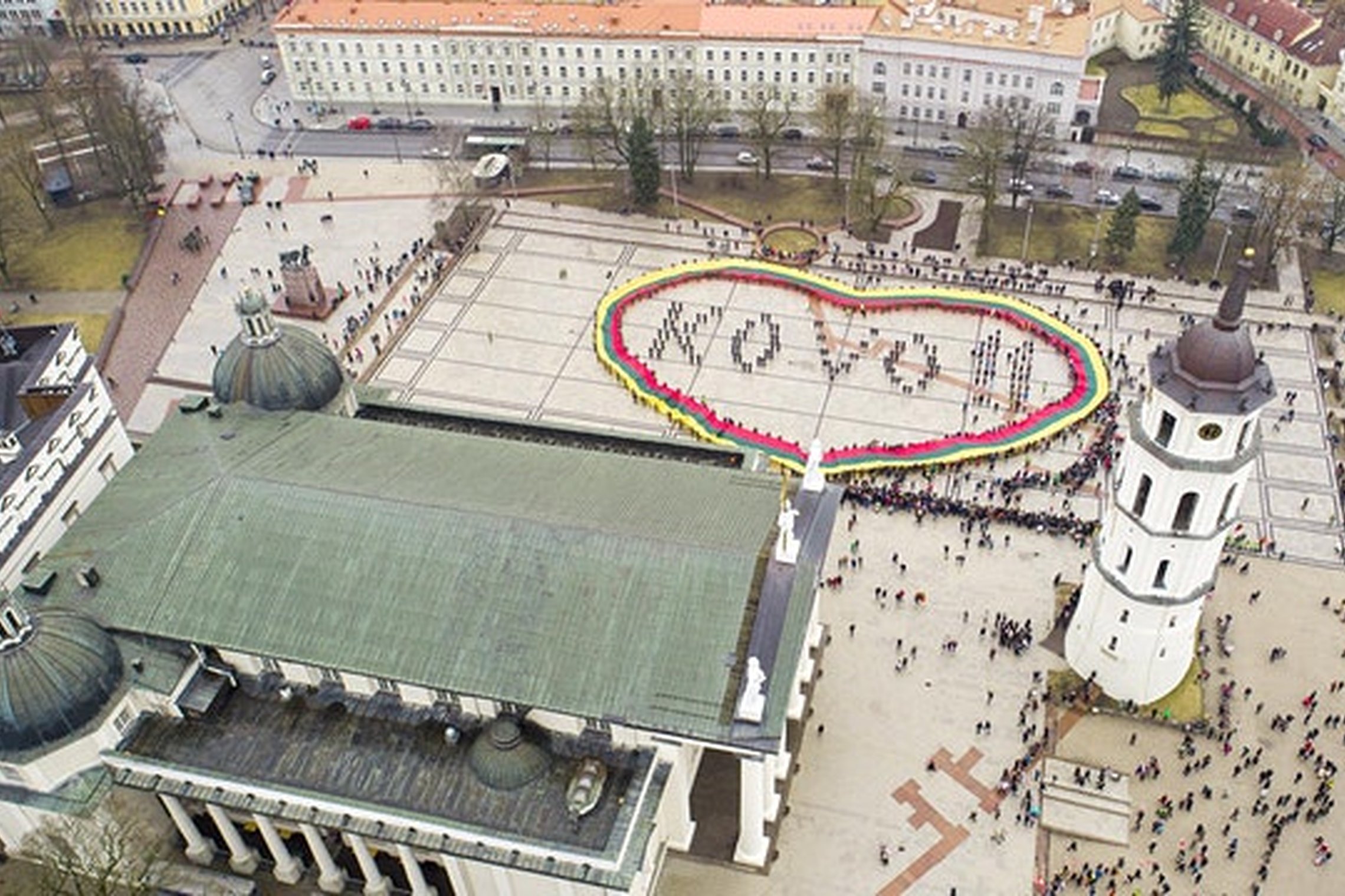Nuotraukoje daryta iš paukščio skrydžio. Tai Vilniaus katedros aikštės nuotrauka. Viršutinėje nuotraukos dalyje yra įvairios įstaigos, apatinėje kairėje pusėje yra katedra, o dešinėje pusėje katedros varpinė. Aikštėje gan daug žmonių, o jos viduryje žmonė