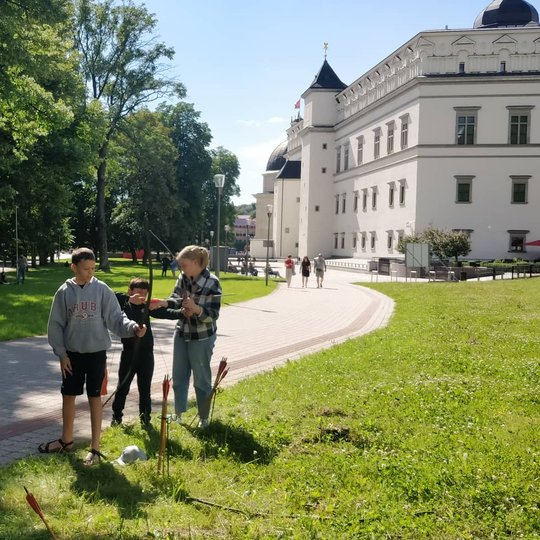 Viduramžiškai apsirengęs vaikinas susirinkusiems žmonėms pasakoja apie lankus. Du berniukai mokomi šaudyti iš lanko.