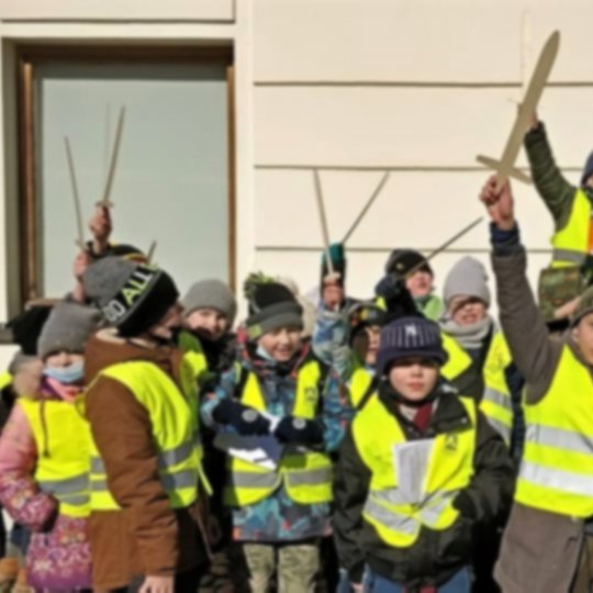 Vaikai, su atšvatinėmis liemenėmis, laiko medinius kalavijus iškėlę į viršų.