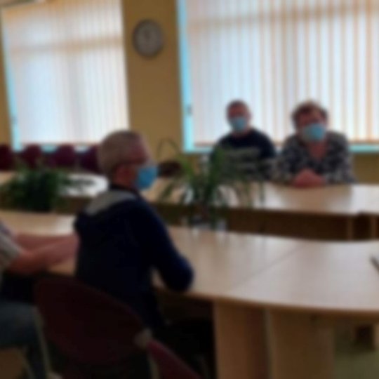 Šiaulių miesto Dienos centro „Goda“ lankytojai sėdėdami prie išilginių stalų klausosi paskaitos nuotoliniu būdu. Lankytojai su medicininėmis kaukėmis.