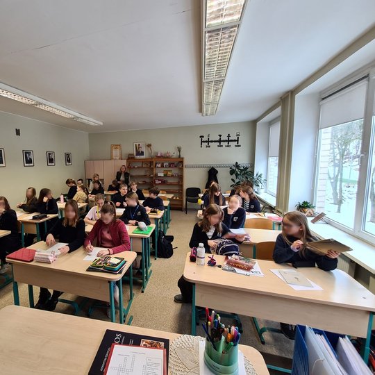 Penktokų klasė, sėdintys suoluose. Klasės gale mokytoja stebi vedamą istorijos pamoką.