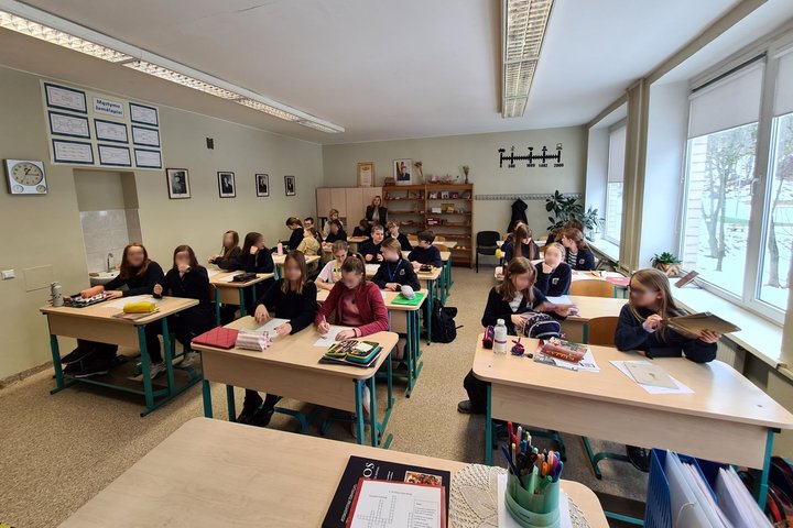 Penktokų klasė, sėdintys suoluose. Klasės gale mokytoja stebi vedamą istorijos pamoką.