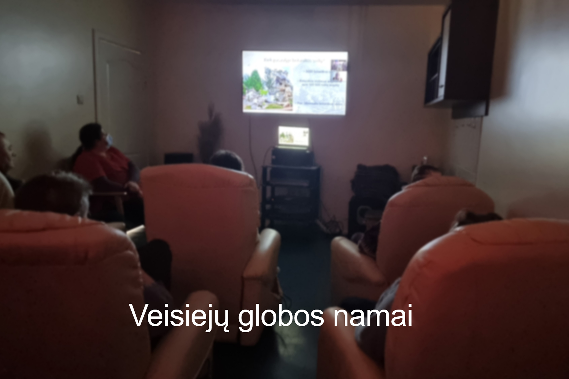 Veisiejų globos namų gyventojai žiūri į ekraną. Nuotraukos apačioje įstaigos pavadinimas.