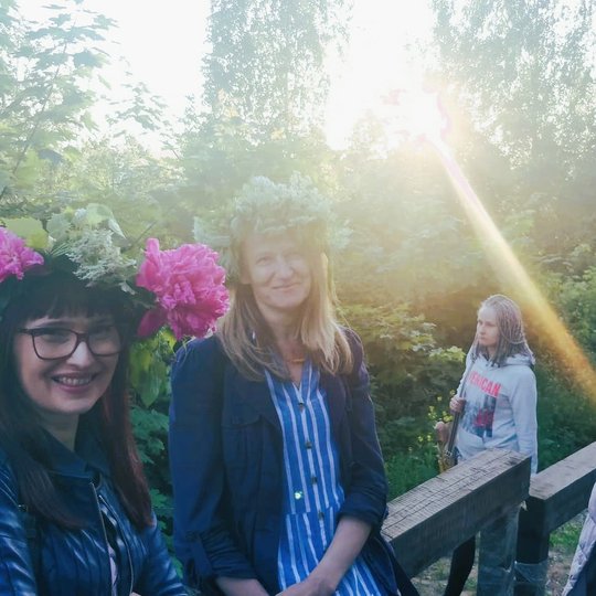 Moterys su šypsenomis ir žolių vainikais atsirėmusios į turėklą pozuoja. Matosi ryškus saulės spindulys.