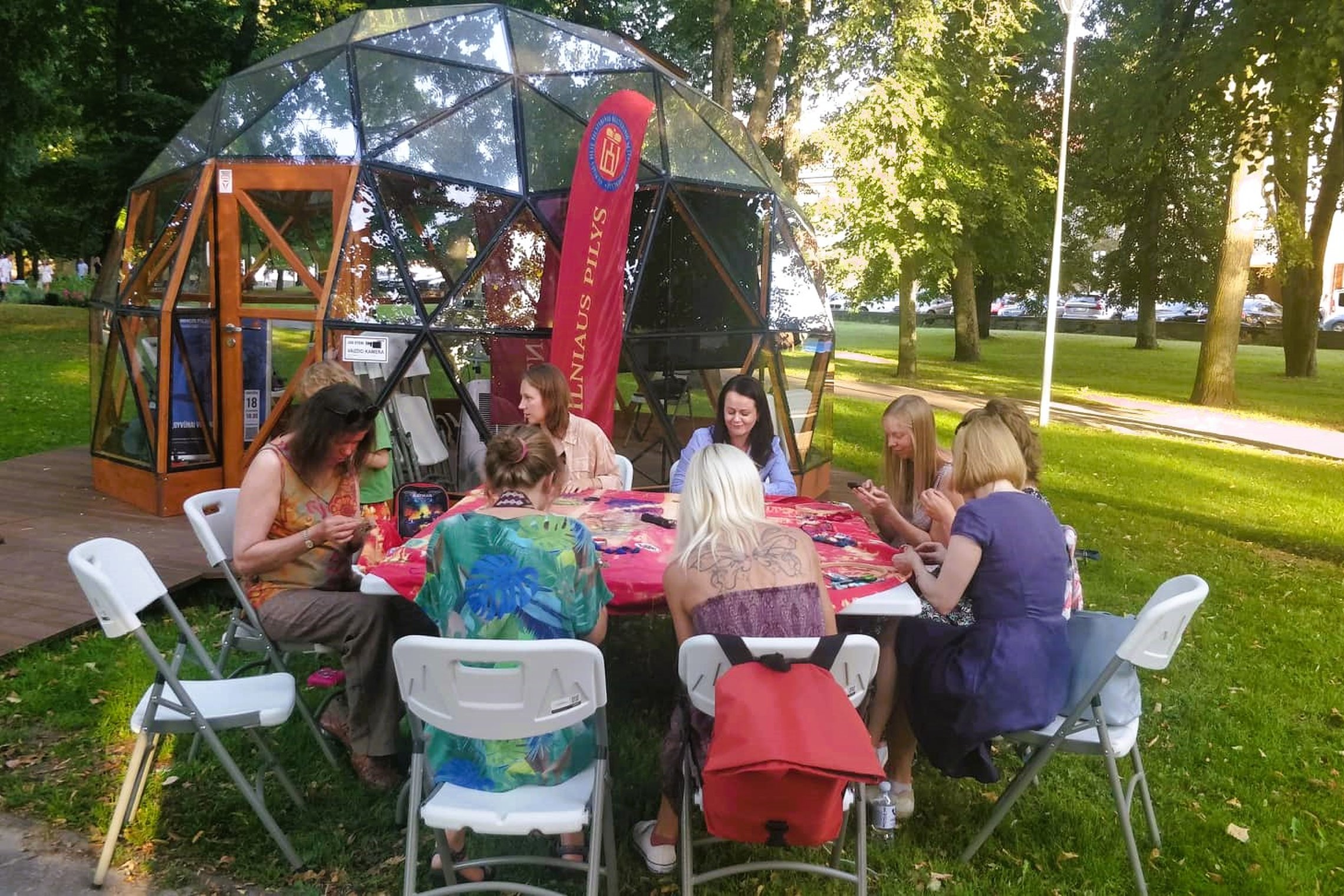 Moteris susėdusios ratu prie balto stalo, siuvinėja audinį lauke, šalia Vilniaus pilių kupolo.