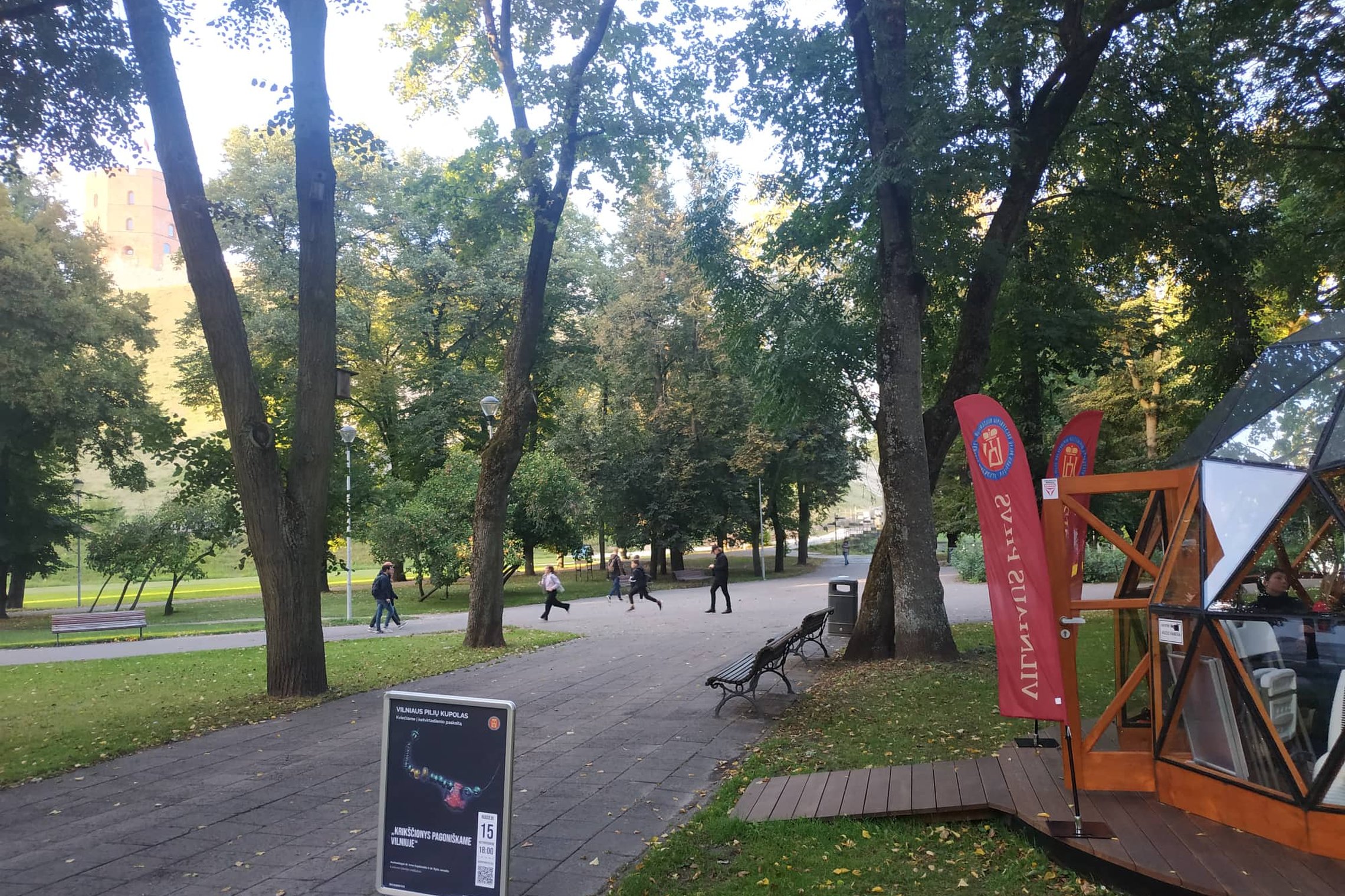 Kairėje Vilniaus pilių kupolas, priešais matosi stendas su paskaitos afiša, dešinėje matosi Gedimino kalnas.