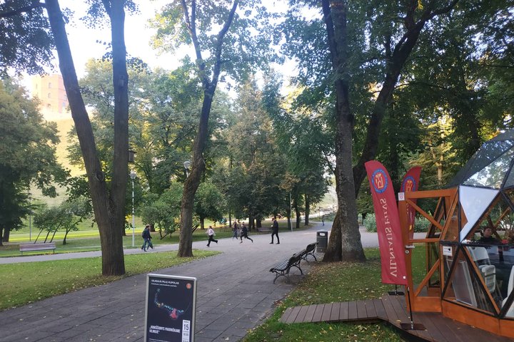 Kairėje Vilniaus pilių kupolas, priešais matosi stendas su paskaitos afiša, dešinėje matosi Gedimino kalnas.