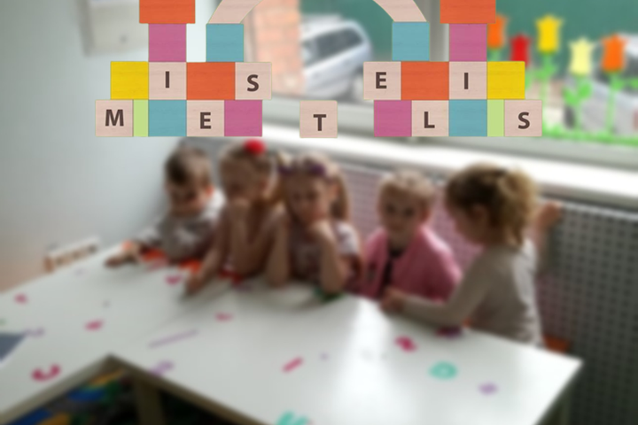 Darželinukai sėdi prie stalo klasėje. Nuotraukos priekyje darželio „Kaladėlių miestelis" logotipas.