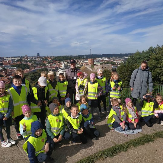 Antrokai mokiniai pozuoja nuotraukai priešais Trijų Kryžių paminklą. Jiems už nugarų Vilniaus senamiesčio panorama.
