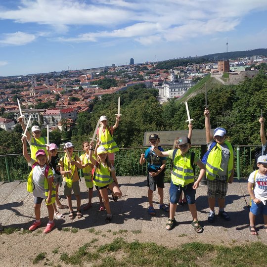 Vaikučiai, medinius kalavijus iškėlę į dangų, pozuoja nuotraukai ant trijų Kryžių kalno. Tolumoje matosi Vilniaus panorama su Gedimino pilim. 