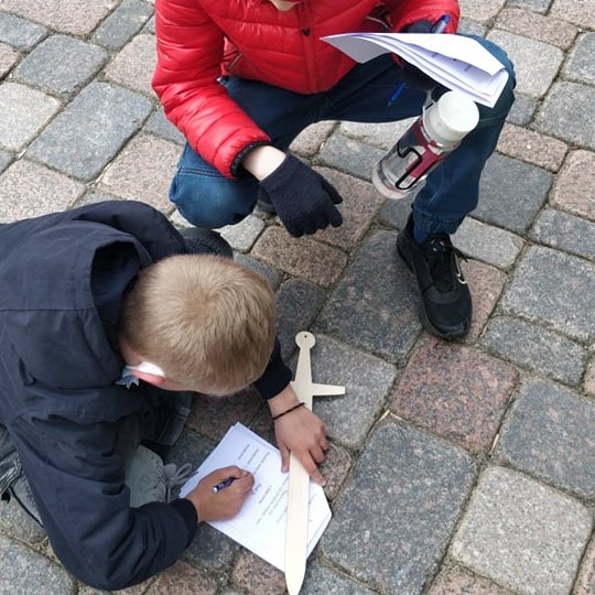 Berniukai, pasidėję medinį kalaviją ant plytelių, pildo orientacinės užduotis.