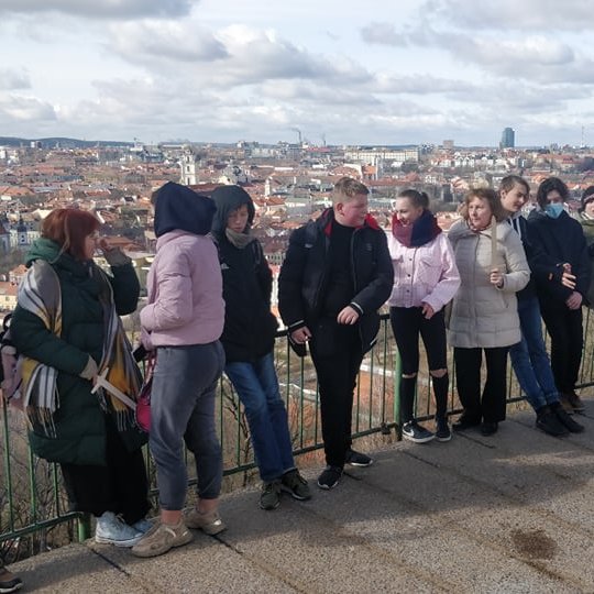 Mokiniai ir mokytojai su mediniais kalavijais, priešais Tris Kryžius. Jiems už nugaros matosi Vilniaus panorama su Gedimino kalnu.