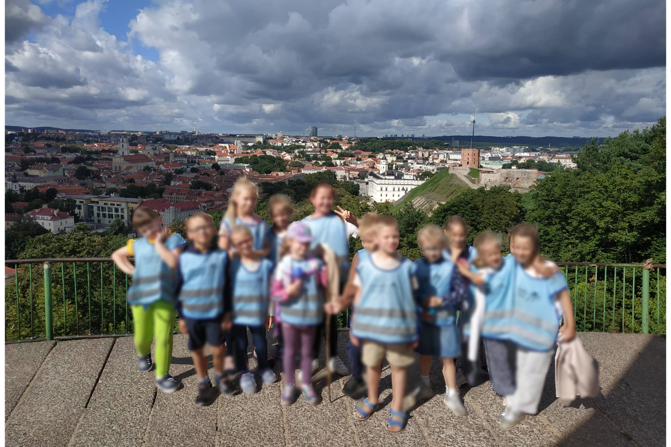 Vaikai su mėlynomis liemenėmis pozuoja nuotraukai. Jiems už nugarų Vilniaus panorama nuo Trijų Kryžių kalno. 
