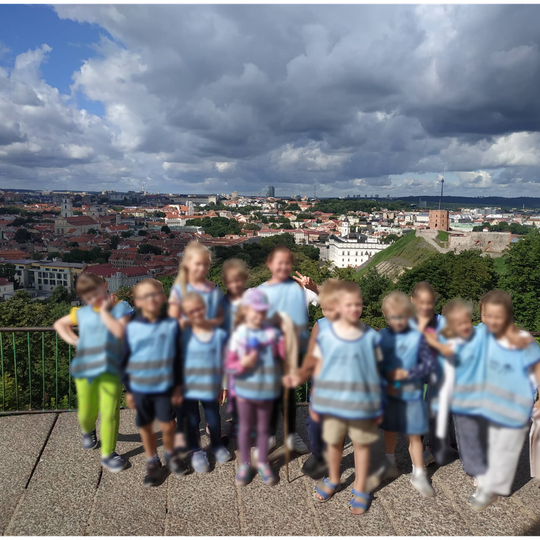Vaikai su mėlynomis liemenėmis pozuoja nuotraukai. Jiems už nugarų Vilniaus panorama nuo Trijų Kryžių kalno. 