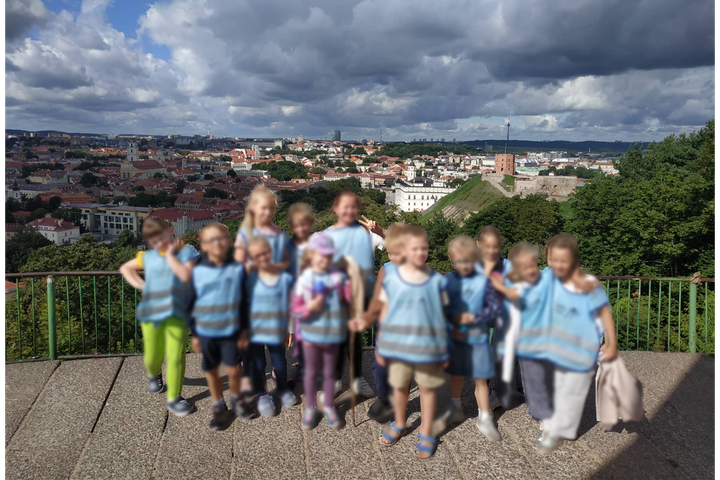 Vaikai su mėlynomis liemenėmis pozuoja nuotraukai. Jiems už nugarų Vilniaus panorama nuo Trijų Kryžių kalno. 