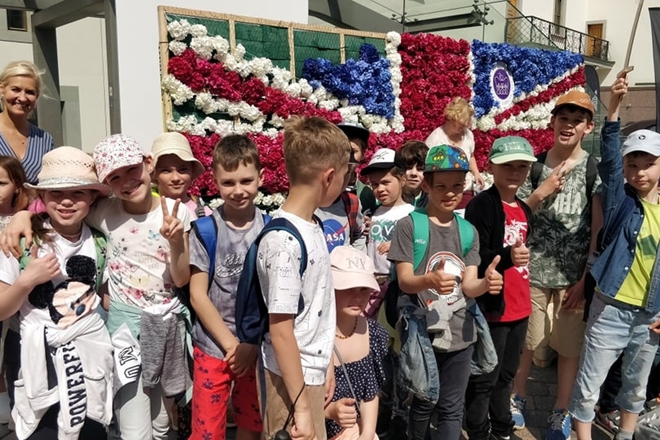 Antrokai kartu su mokytoja pozuoja bendrai nuotraukai. Jiems už nugarų iš gėlių puokščių sudaryta Jungtinės Karalystės vėliava. 