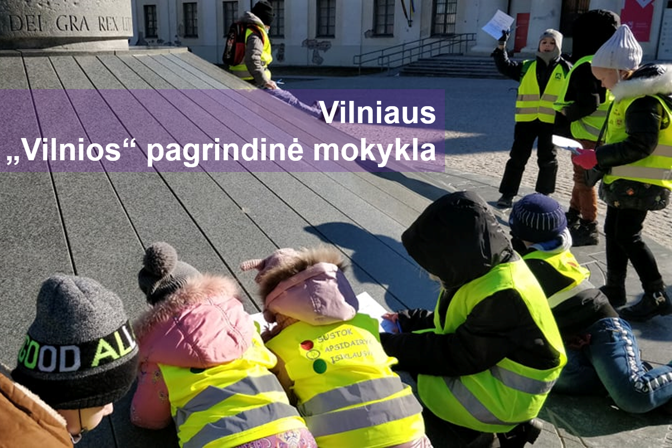 Pradinukai pildo užduotis šalia karaliaus Mindaugo paminklo. Nuotraukos priekyje parašyta "Vilniaus „Vilnios“ pagrindinės mokykla".