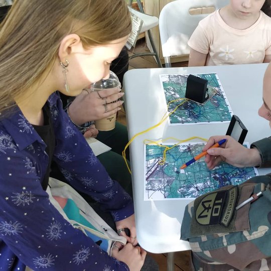 Šaulė su tušinuku rodo tašką žemėlapyje, aplinkiniai klausosi jos. 