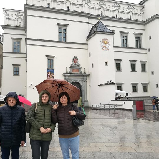 Lietingą diena, tris žmonės pozuoja nuotraukai priešais Valdovų rūmus.