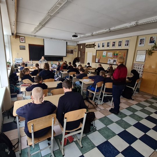 Mokiniai klausosi mokytojos, stovinčios klasės gale. Mokiniai sėdi suoluose.