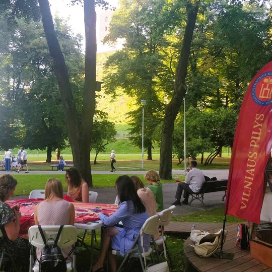 Šalia Vilniaus pilių kupolo, lauke, moteris ratu susėdusios šalia stalo, siuvinėją bendrą audinį ir kalba apie tvarumą.