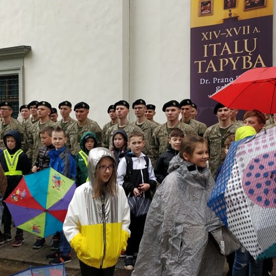 Vaikai su lietpalčiais ir skečiasi, o už jų išsirikiavę uniformuoti kareiviai iš inžinerijos bataliono.