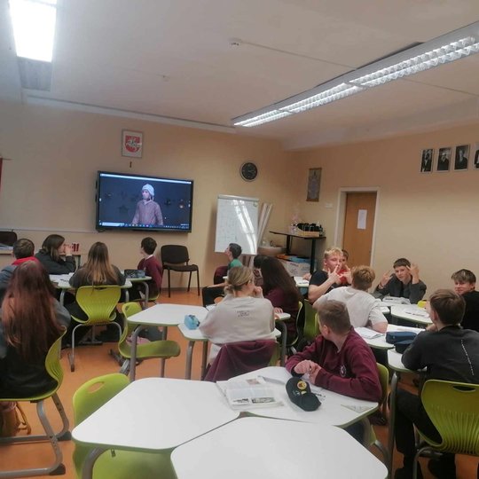 Klasėje mokiniai klausosi pamokos ir žiūri į skaidres per projektorių.
