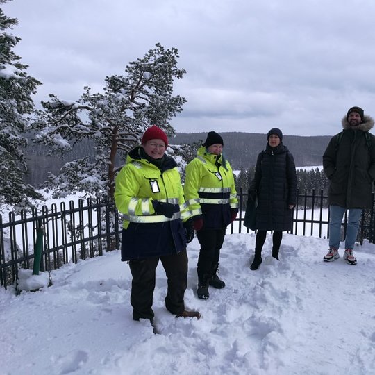 Rezervato darbuotojai kartu su kolegomis iš Suomijos pozuoja nuotraukai. Jiems už nugarų apsnigtų mišku peizažas.
