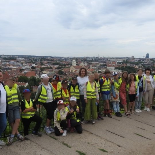 Devyniolika smalsučių ir trys mokytojos pakiliomis nuotaikomis pozuoja priešais Vilniaus senamiesčio panoramą.