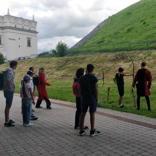 Viduramžiškai apsirengęs vaikinas prižiūri kaip du berniukai šaudo iš lankų. Jiems už nugarų žiūri praeiviai.
