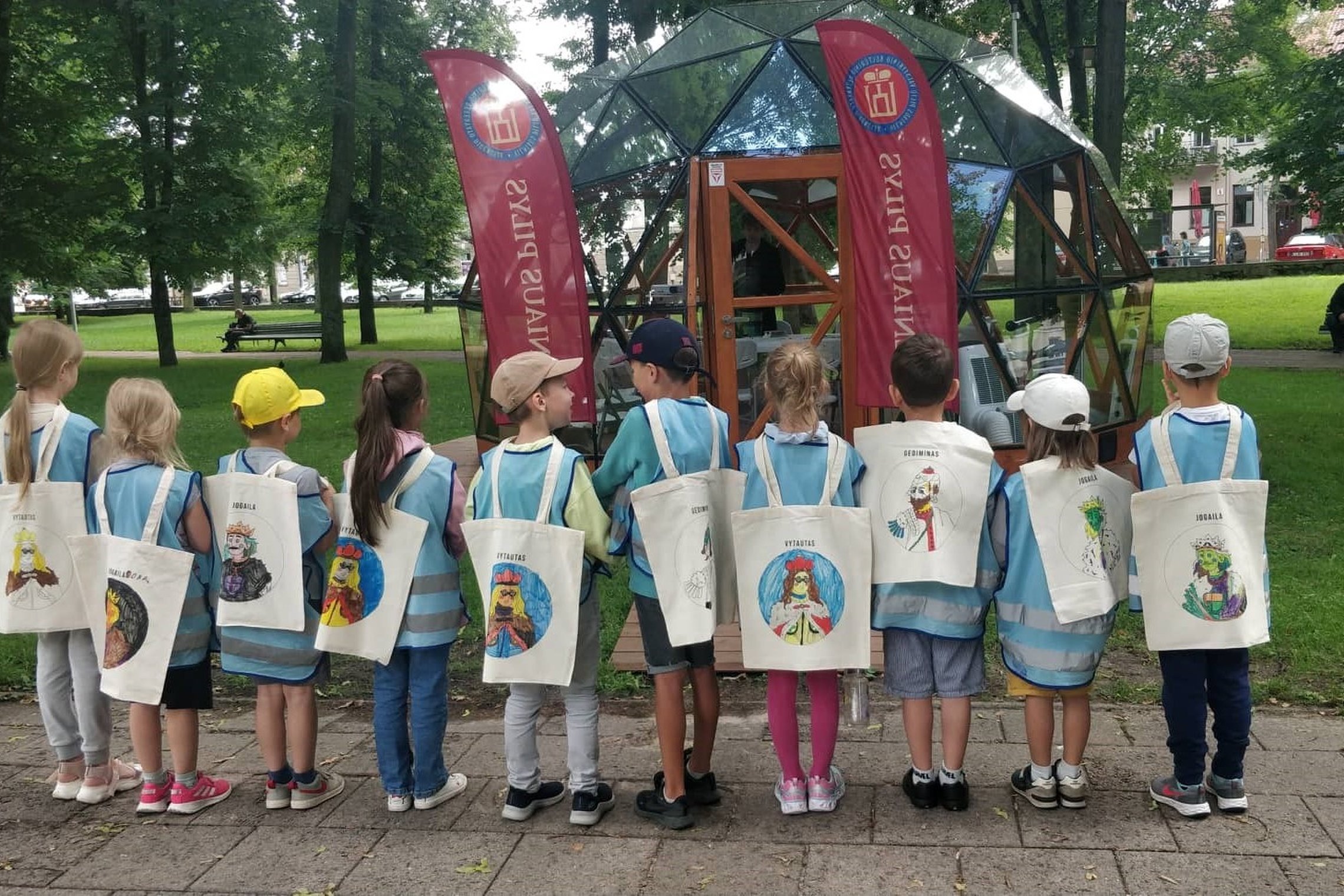 Vaikai pozuoja atsistoję veidu  prieš Vilniaus pilių kupolą,  ant nugaros užsidėję medžiaginius maišelius su nuspalvintais Jogailos, Gedimino ir Vytauto atvaizdais. 