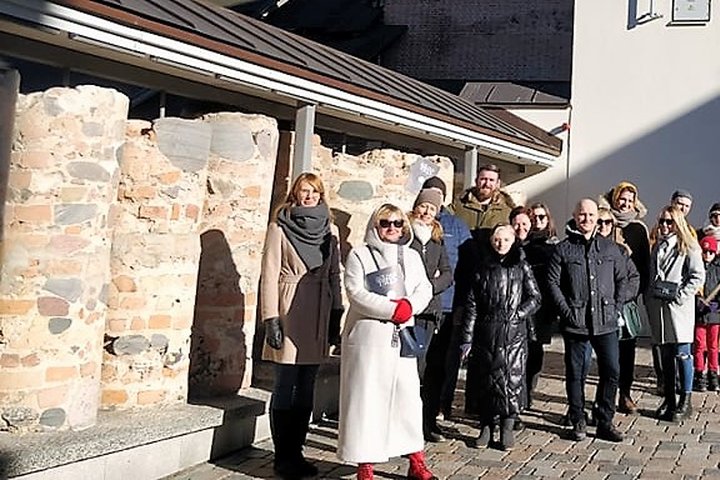 Būrelis žmonių šalia eksponuojamų Žemutinės pilies autentiškų mūro liekanų. Vienas iš Valdovų rūmų kiemelių, kuriame, po dailiu stogeliu eksponuojama poros metrų, didelių akmenų bei plytų mūro siena, statmenai prisišliejusi prie akinančiai baltos sienos v