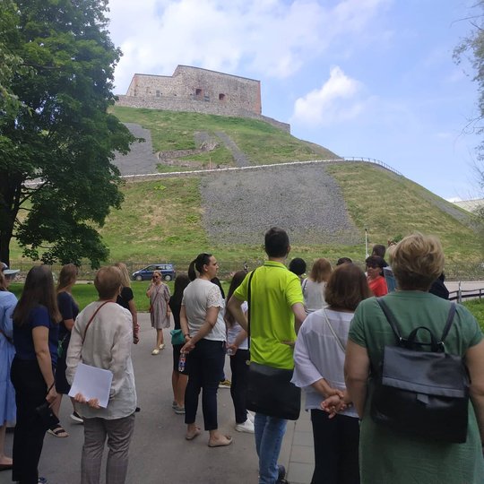 Žmonės klausosi ekskursijos Trispalvės alėjoje. Fone matosi rytinę Gedimino kalno pusė. 