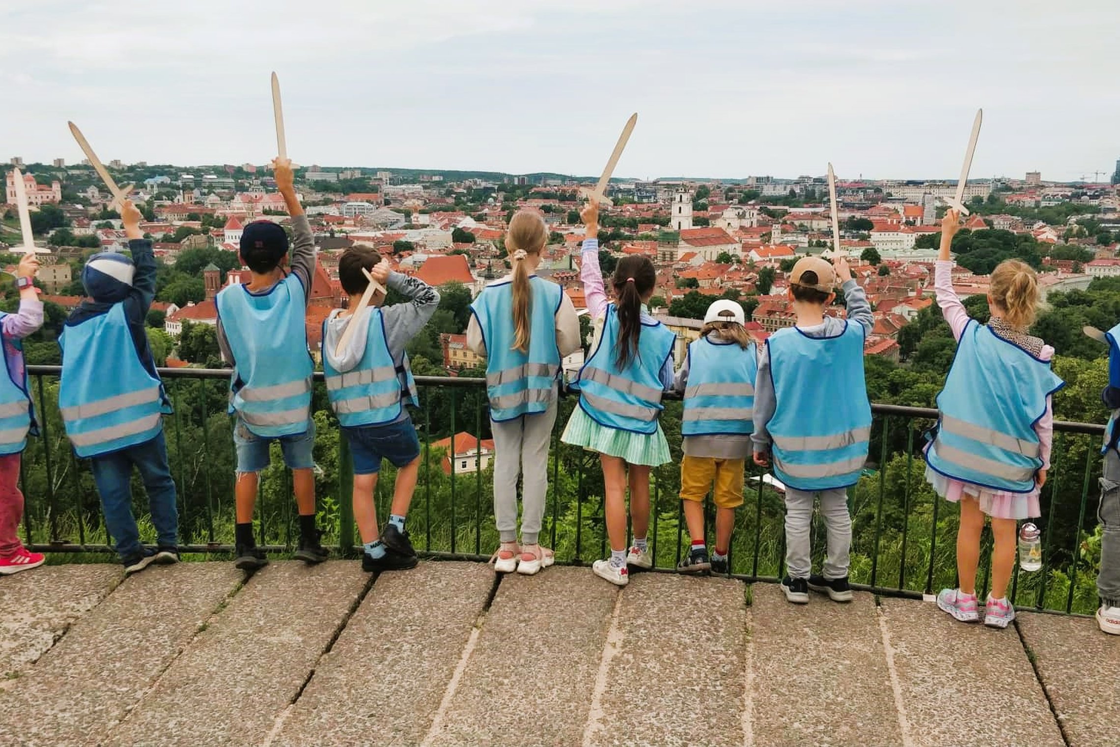 Vaikai su mėlynomis  atšvaitinėmis liemenėmis iškėlę medinius kalavijus žiūri nuo Trijų kryžių kalno. 