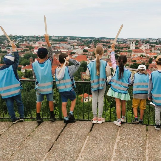 Vaikai su mėlynomis  atšvaitinėmis liemenėmis iškėlę medinius kalavijus žiūri nuo Trijų kryžių kalno. 