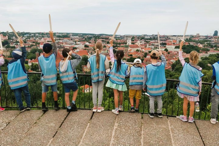 Vaikai su mėlynomis  atšvaitinėmis liemenėmis iškėlę medinius kalavijus žiūri nuo Trijų kryžių kalno. 