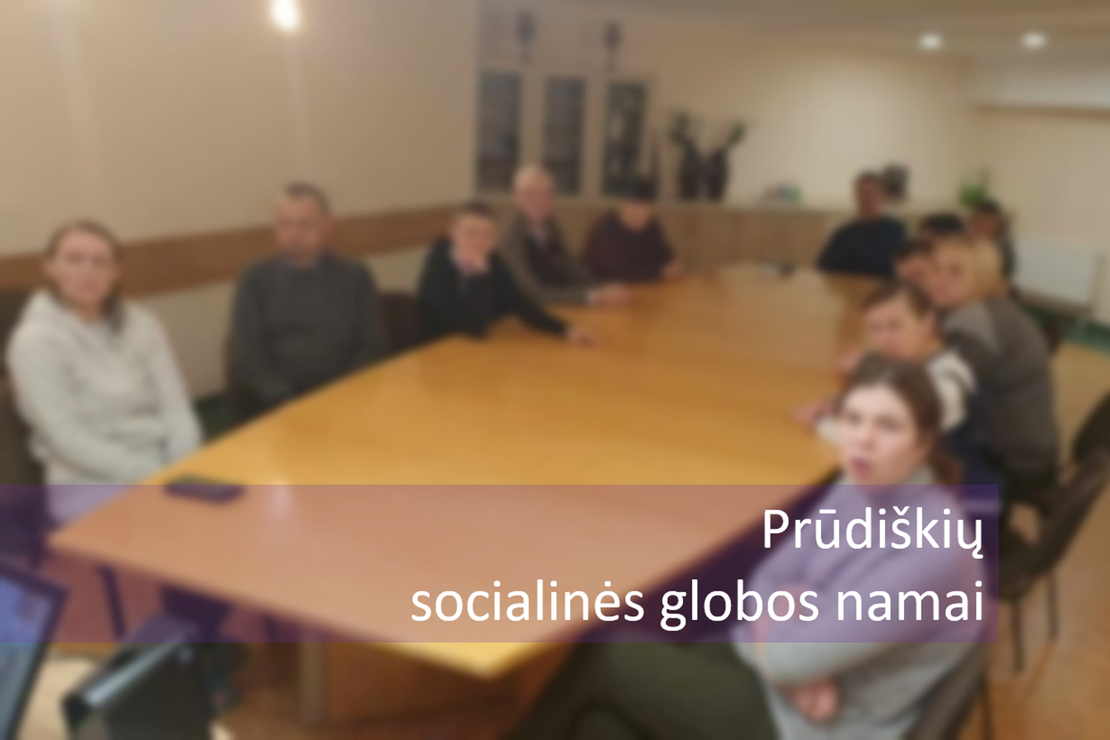 Žmonės sėdi prie pailgo medinio stalo, kambaryje, Nuotraukos priekyje parašyta "Prūdiškių socialinės globos namai".