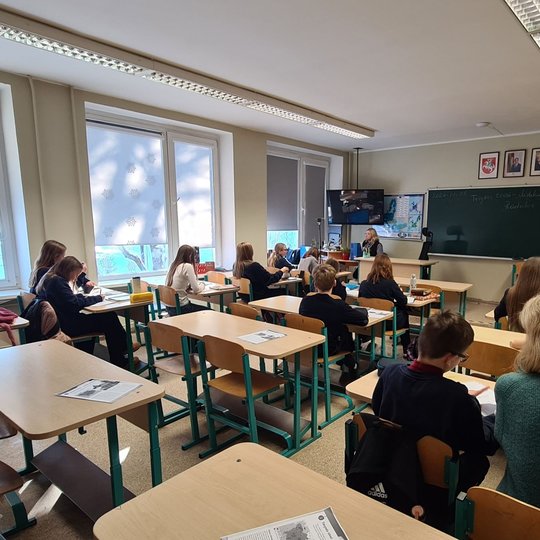 Mokiniai klausosi istorijos pamokos. Mokytoja sėdi prie stalo.