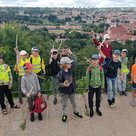 Vaikai, iškėlę medinius kalavijus į viršų, pozuoja nuotraukai. Jiems už nugaros Vilniaus panorama nuo Trijų Kryžių.