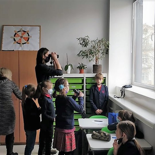 Vaikai su mokytojomis ir ornitologu Gediminu Petkumi žiūri per langą, naudodamiesi žiūronus.