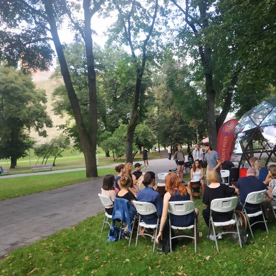 Užsiėmimo dalyviai ant kėdžių, susėdę ratu, muša būgnus šalia Vilniaus pilių kupolo.