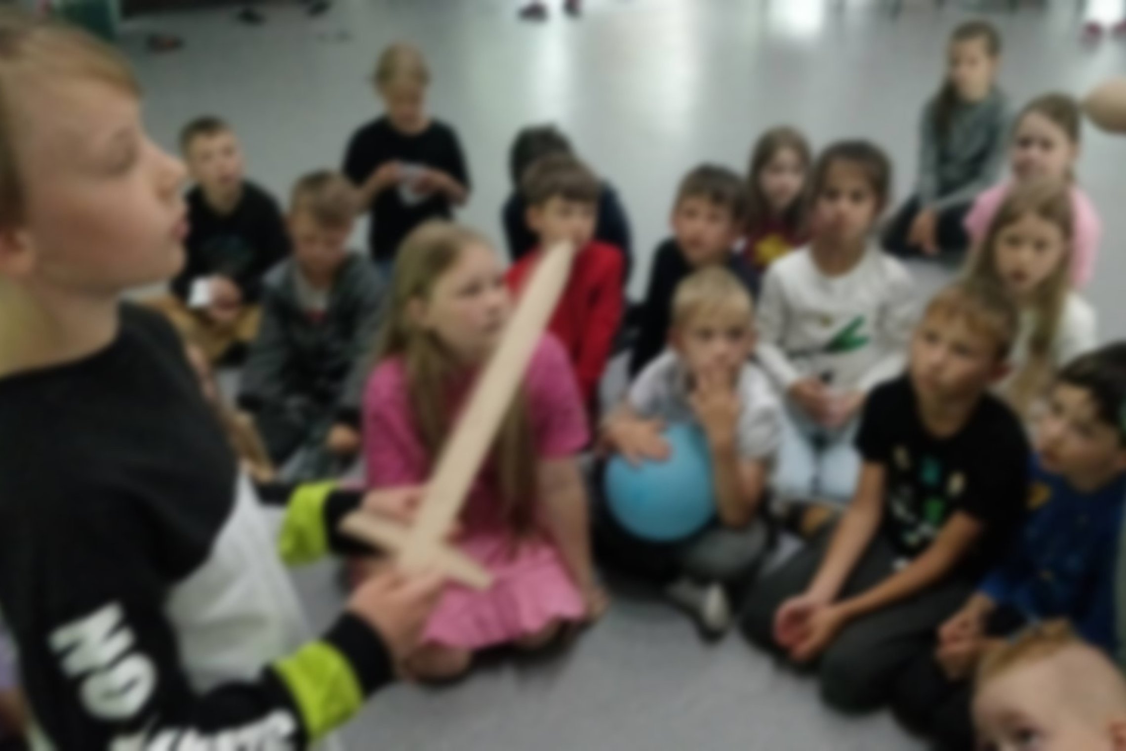 Vaikai atsiklaupę klauso edukacijos. Berniukas priekyje laiko medinį kalaviją.