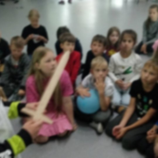 Vaikai atsiklaupę klauso edukacijos. Berniukas priekyje laiko medinį kalaviją.
