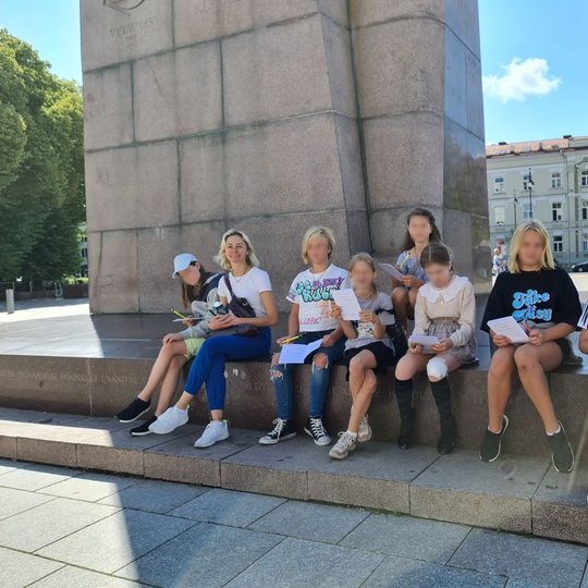 Ekskursantai, prisėdę į pavėsį prie kunigaikščio Gedimino paminklo.